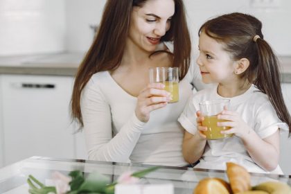 Happy mother and daughter in casual clothes drinking juice and looking at each other while having breakfast together in contemporary kitchen