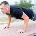 Adult male exercising outdoors performing push-ups with earphones and water bottle.