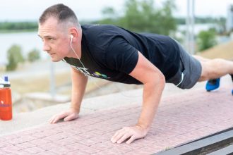 Adult male exercising outdoors performing push-ups with earphones and water bottle.